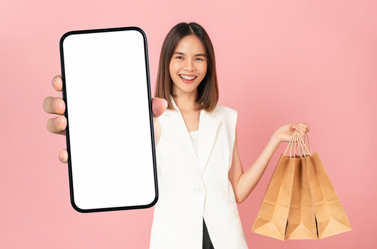 Beautiful Asian woman holding brown blank craft paper shopping bags and smartphone on pink background.