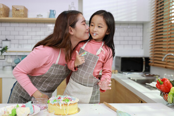Happy lovely asian single mom and cute daughter in apron having fun with decorating homemade cake in the kitchen. Mother kiss daughter cheek. Family lifestyle cooking with education concept.