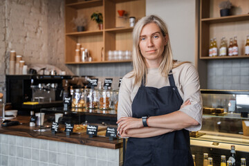 Portrait of aged woman barman dressed in apron posing with crossed arms in coffee shop.