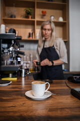 Shot of coffee cup on table and aged female barista with blond hairs.