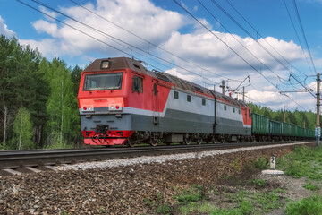 A red electric locomotive with a freight train moves along the railroad tracks.