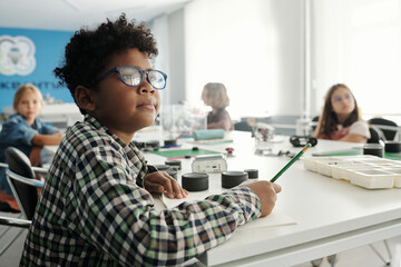 Obraz premium Clever schoolboy in eyeglasses and checkered shirt making notes in copybook while sitting by table against his classmates