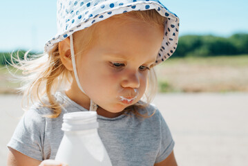 Funny portrait of child drinking milk, pouting cheeks. Little blonde girl, toddler in hat with...