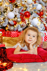 a charming little girl curly blonde in a white dress lies on the floor on a pillow and looks at the picture against the background of Christmas trees in the bright interior of the house