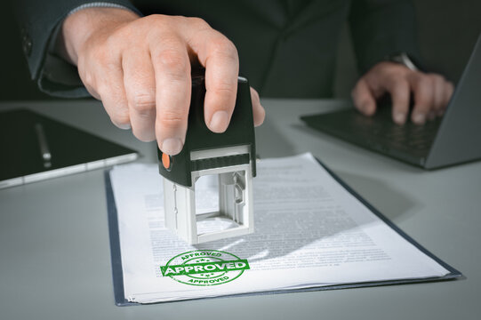 Hands of businessman stamp on paper document to approve business investment contract agreement. Close-up Of Person Hand Stamping With Approved Stamp On Document At Desk.