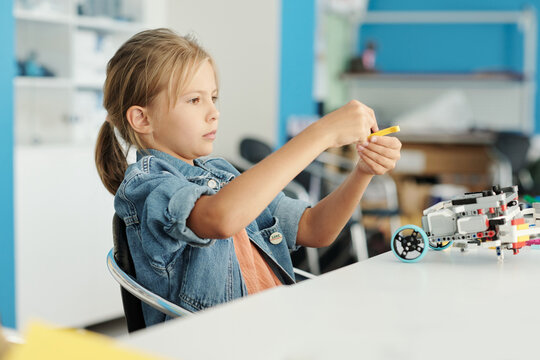 Cute little girl in casualwear connecting details of electronic toy while sitting by desk at lesson of robotics and creating new robot