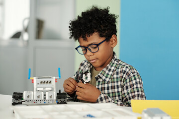 Clever little boy in casualwear and eyeglasses connecting details of toy robot while sitting by...