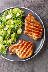 Grilled pork steak and green vegetable salad closeup in the plate on the table. Vertical top view from above