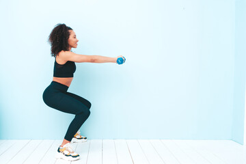 Fototapeta na wymiar Fitness smiling black woman in sports clothing with afro curls hairstyle.She wearing sportswear. Young beautiful model doing squats.Female holding dumbbells in studio near light blue wall