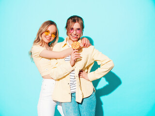 Two young beautiful blond smiling hipster female in trendy summer clothes. Sexy carefree women posing near blue wall in studio. Trendy and positive models having fun with candy sweet lollipop