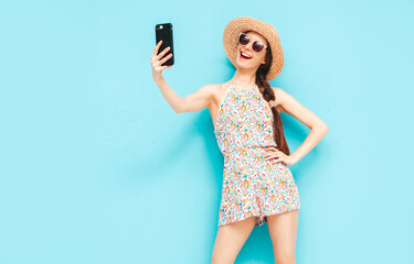 Portrait of young beautiful smiling female in trendy summer overalls. carefree woman posing near blue wall in studio. Positive model having fun indoors. Cheerful and happy. In hat. Takes selfie