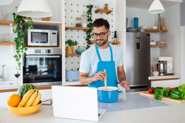 A young man spends a day at home, prepares food of vegetables in the kitchen, a man in home clothes and with a beard, uses a laptop to learn online cooking