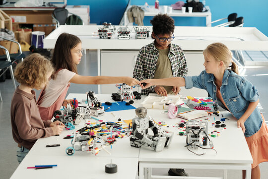 Group of youthful intercultural schoolkids constructing new robots at lesson while helpful schoolgirl passing detail to classmate