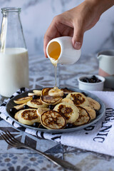A woman's hand pours honey on mini pancakes with bananas and berries. The background is light, vertical orientation, side view.