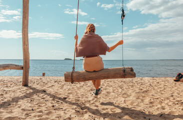 A beautiful woman is swinging on a swing overlooking the sea. Autumn at sea. Sand, water, swing. Rest by the water.