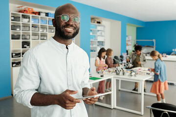 Happy young black man with tablet standing in front of camera against group of intercultural schoolkids creating new robots