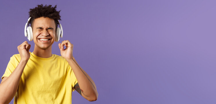 Portrait of pleased, excited young man enjoying nice quality awesome beats in headphones, close eyes and smiling rejoicing, dancing over cool new song, listen music over purple background