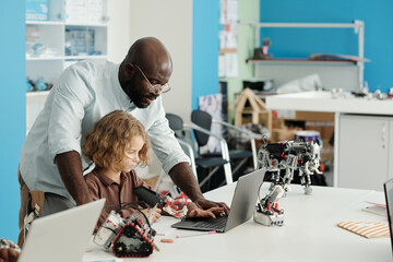 Young African American teacher of robotics bending over cute schoolboy sitting by desk in front of laptop and working over new project