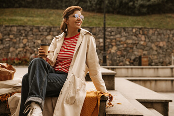 Pretty young caucasian woman in casual clothes with coffee sitting on steps in city park. Short-haired beauty in transparent glasses looks to side. Concept of spending time alone