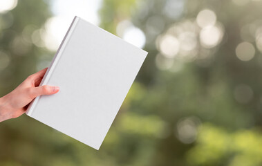 Hand holding book mockup on blurred background with green trees, grass. Literature about nature, ecology. Reading leisure, harmony concept