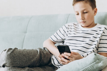 Teenage boy sitting on couch using smartphone and playing video games on internet online at cozy home. Child holding mobile phone and looking at screen. Social media
