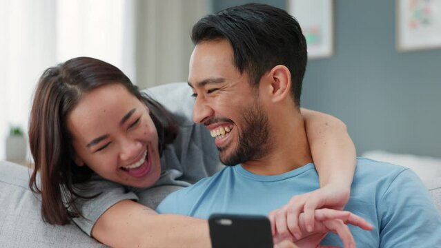 Asian couple, phone or laughing in hug on sofa in house living room or home interior. Happy smile, comic or fun bond man or woman or playful boyfriend or girlfriend with mobile tech for internet game