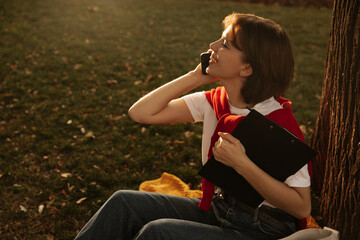 Side view of young caucasian woman talking on smartphone with smile on her face outdoors. Short-haired girl holds notebook in her hands. Cell phone use concept