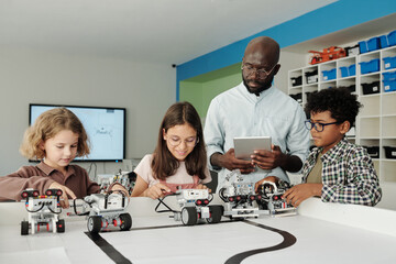 Young confident African American teacher of robotics with tablet helping cute intercultural schoolkids control their robots during play