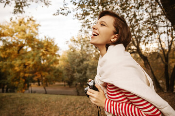 Cheerful young caucasian lady laughs from what she saw, holds camera in her hands outdoors. Brunette girl has fun alone. Rest and recovery concept.