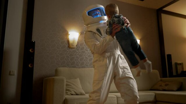 A male father in an astronaut costume begins to dance with toddler's son in the interior of a cozy apartment. The concept of initiating a child into his interesting work in space on a spaceship.