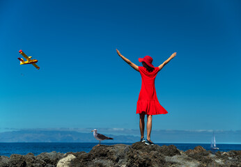 Happy woman in red standing on the rocky ocean shore with open hands. Calm ocean water and pure blue sky at the background.