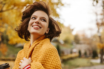 Attractive young caucasian girl with flying short hair looking to side in street. Model with perfect smile wears yellow scarf. Rest time concept.