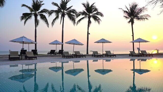 A pink and yellow tropical sunset behind a row of palm trees, empty poolside lounge chairs, and beach umbrellas.