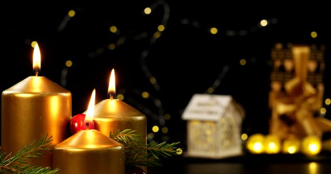 Three golden Christmas candles with fir branches and red berries burn in the dark. In the background of candles, a blurred Christmas picture is a small house a gift and a garland.
