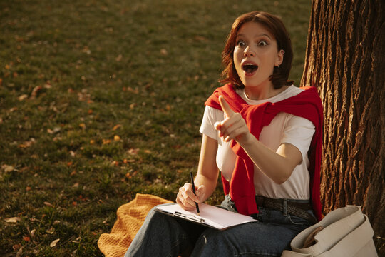 Inspired Young Caucasian Woman Caught Idea With Mouth Wide Open While Sitting Outdoors On Warm Day. Brunette Makes Sketches Of Project. Technologies And Concept Of Remote Work.