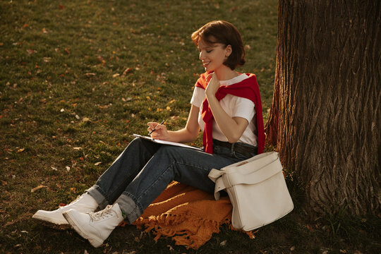 Pretty young caucasian woman is sitting on grass near tree painting landscape of autumn park. Short-haired girl crippled by hobby. Modern education, student body concept.