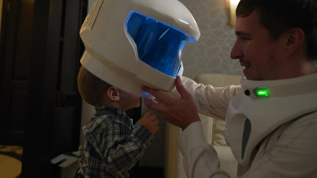 The astronaut father gives his son a helmet from the suit to try on. A man plays with a boy and puts on a white astronaut helmet with blue lighting for playing in the interior of the apartment.
