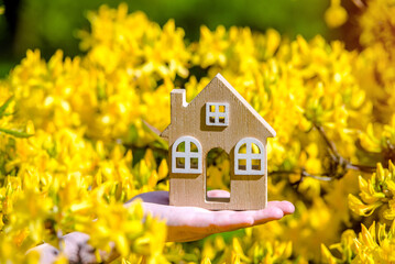 The girl holds the house symbol against the background of blossoming yellow rhododendron