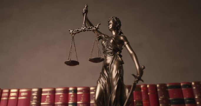 concept of justice illustrated by bronze blindfolded goddess holding sword and scale, rotating and moving in front of row made of books in a university room