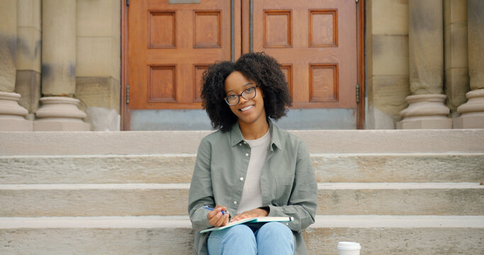 African charming young lady with curly hair and glasses, smiling toothily seats on the stairs outside and writing in her notepad