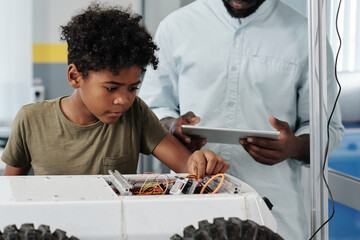 Cute African American schoolboy connecting cables of toy robot while following instructions of his...