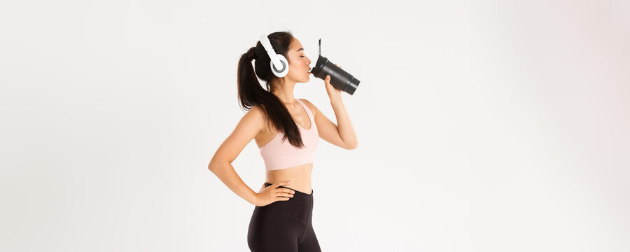 Sport, wellbeing and active lifestyle concept. Profile portrait of sexy asian fitness coach, woman in headphones drinking water from bottle during workout in gym, standing white background