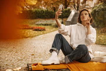 Cute fair-skinned young girl makes selfie on smartphone showing two fingers while sitting on park bench. Brunette is wearing white hoodie, jeans and boots. Autumn trendy street style, concept