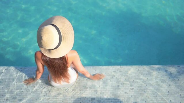Back to the camera, a woman in a large straw sun hat sits on the step of a swimming pool. 