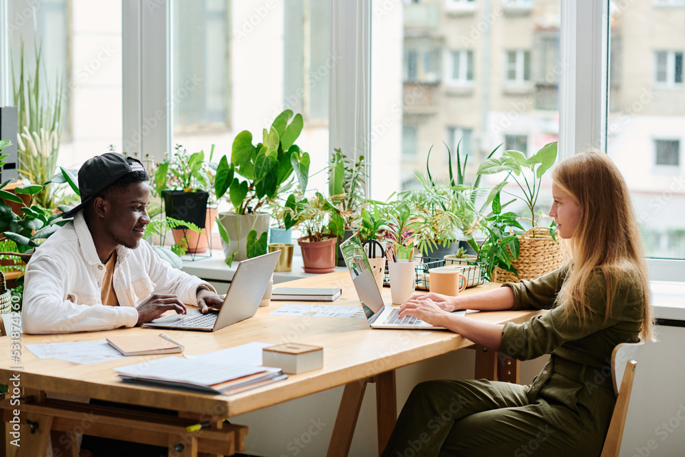 Poster two young multicultural coworkers sitting by desk in front of laptops and networking against window 
