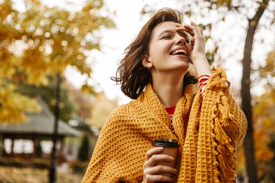 Funny young fair-skinned brown-haired lady laughs with her head tilted in fresh air. Girl wrapped yellow scarf, holds paper cup with coffee. Cozy atmosphere in park, autumn concept.