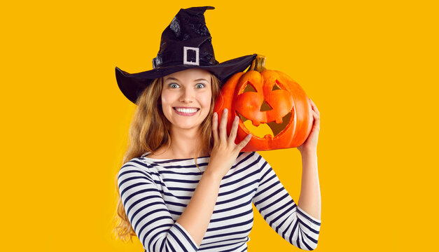 Portrait of a happy woman with a pumpkin. Beautiful young girl in a witch hat standing isolated on a yellow background, holding an orange pumpkin, looking at the camera and smiling. Halloween concept