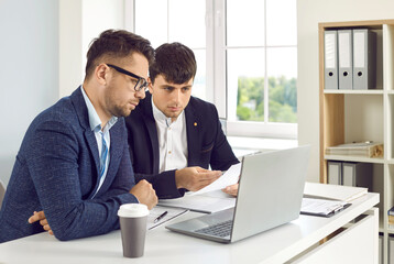 Business people working together in office. Two serious men in suits sitting at desk with laptop computer and discussing project. They are working with documents and looking at laptop screen.