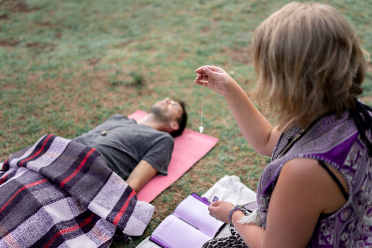 Unrecognizable woman with amulet doing hypnosis to calm lying man in nature