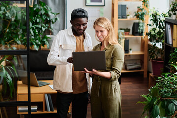 Two young intercultural employees in casualwear looking at screen of laptop held by blond...